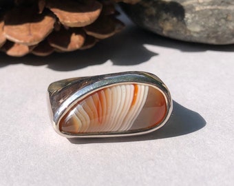 May include: A silver ring with a white and orange agate stone set in the center. The stone has a unique pattern of white and orange stripes.