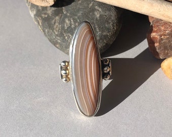 May include: A silver ring with a brown and white agate stone set in the center. The stone has a unique striped pattern. The ring is accented with small silver beads on either side of the stone.