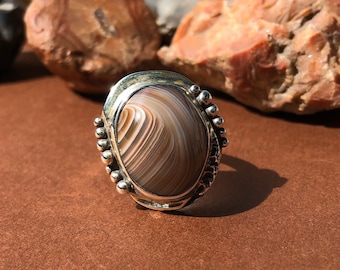 May include: A silver ring with a brown and white agate stone set in a silver bezel. The ring is decorated with small silver beads around the bezel.