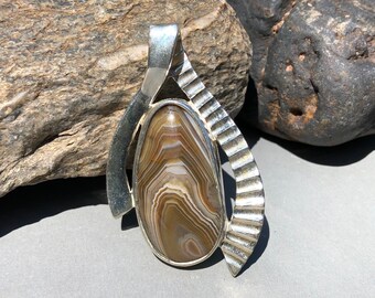 May include: A silver pendant with a large, oval-shaped agate stone. The stone features brown and white banded patterns. The silver setting has a unique, asymmetrical design with a curved, ribbed accent on one side. The pendant is displayed on a gray surface.