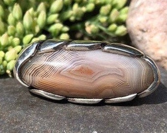 May include: A silver ring with a light brown agate stone set in a silver setting. The stone has a swirling pattern of light and dark brown.