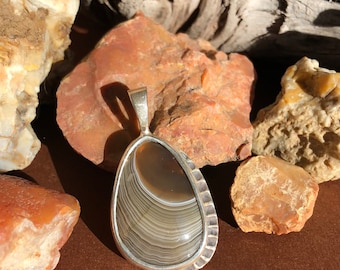 May include: A silver pendant with a brown and white agate stone. The stone has a layered pattern and is set in a silver bezel.