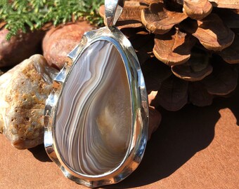 May include: A silver pendant with a white and brown agate stone. The stone has a wavy pattern and is set in a silver frame with a scalloped edge.