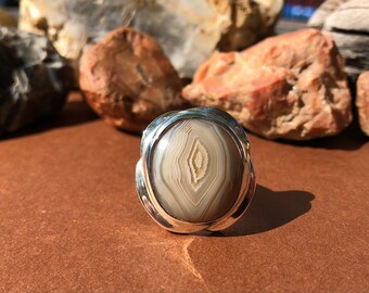 May include: A silver ring with a large oval agate stone. The agate is a light brown color with white and gray banding.