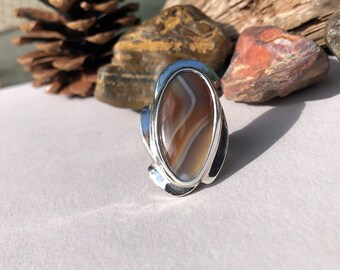 May include: A silver ring with an oval-shaped agate stone. The stone has brown, white, and gray stripes.