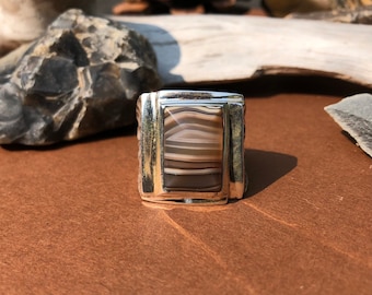 May include: A silver ring with a rectangular brown and white agate stone set in the center. The ring has a simple, modern design.