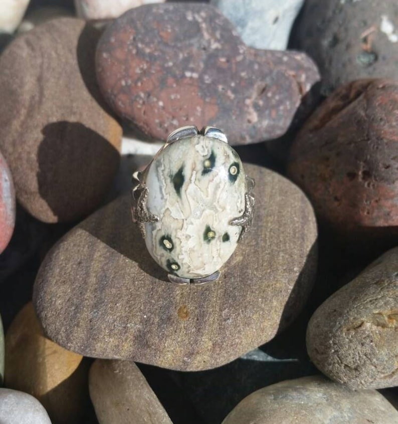 May include: A silver ring featuring a white and green oval stone set in a claw setting. The stone has a distinctive pattern of black and white markings.