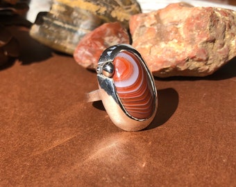 May include: A silver ring with a red and white agate stone set in a bezel setting. The stone has a swirling pattern of red, white, and brown.