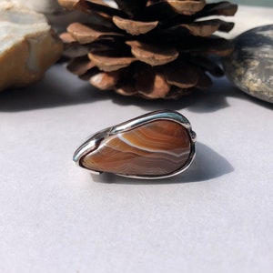 May include: A silver ring with a brown agate stone set in the center. The stone has a swirling pattern of brown and white.