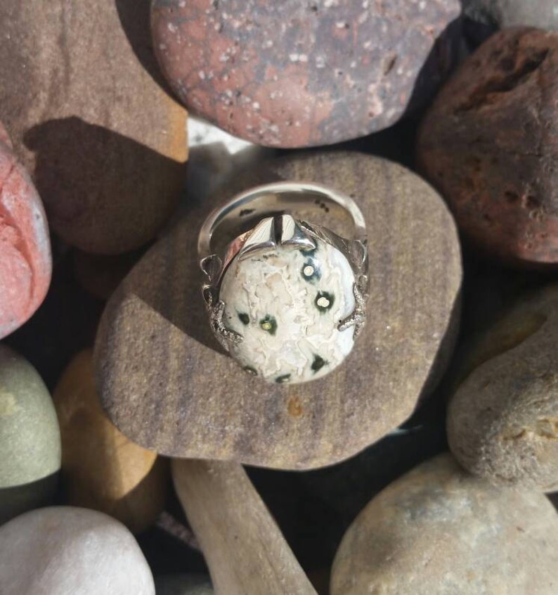 May include: A silver ring featuring a white and green stone set in a silver setting. The ring is surrounded by smaller stones.