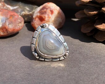 May include: A silver ring with a gray agate stone set in a textured bezel. The stone has a swirling pattern of white and gray.