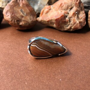 May include: A silver ring with a teardrop-shaped brown agate stone set in a silver bezel. The agate has white and brown bands.