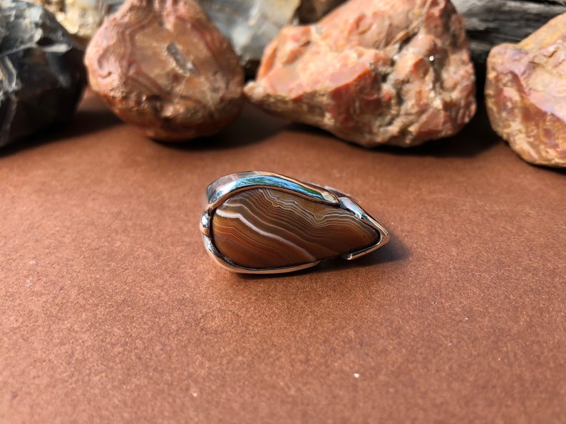 May include: A silver ring with a teardrop-shaped brown agate stone set in a silver bezel. The agate has white and brown bands.