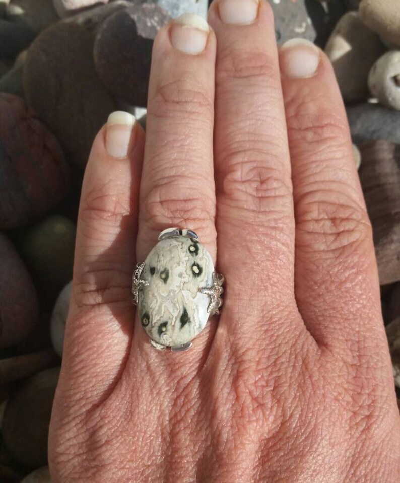 May include: A silver ring featuring a white and black stone set in a silver setting. The stone has a swirling pattern and black spots.