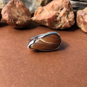 May include: A silver ring with a brown agate stone set in the center. The stone has a swirling pattern of brown and white.