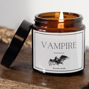 May include: A lit soy wax candle in a brown glass jar with a black lid. The label reads "VAMPIRE" with a bat illustration. The candle sits on a wooden surface.
