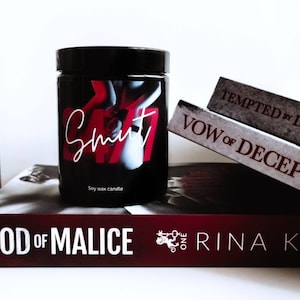 Puede incluir: Una vela negra de cera de soja con la palabra "Smut" escrita en blanco en la parte delantera. La vela est&aacute; sentada sobre una pila de libros con los t&iacute;tulos "God of Malice", "Vow of Decep" y "Tempted by L".