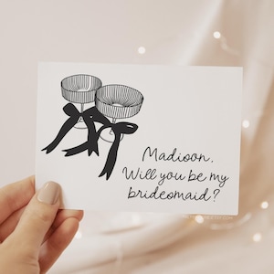 May include: A white card with a black and white illustration of two champagne flutes tied with black ribbons. The text on the card reads "Madison, Will you be my bridesmaid?"