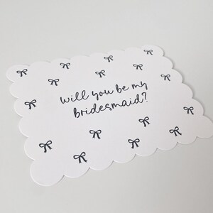 K&ouml;nnte beinhalten: Eine wei&szlig;e Karte mit einem gewellten Rand und dem Text "Will you be my bridesmaid?" in schwarzer Kursivschrift. Die Karte ist mit kleinen schwarzen Schleifen verziert.