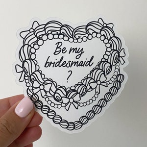 May include: A white heart-shaped sticker with a black outline and a black and white illustration of a necklace with bows. The text "Be my bridesmaid?" is written in black inside the heart.