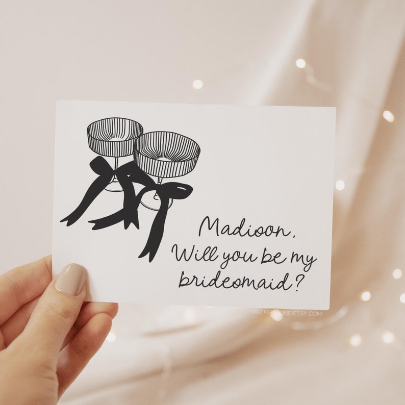 May include: A white card with a black and white illustration of two champagne flutes tied with black ribbons. The text on the card reads "Madison, Will you be my bridesmaid?"