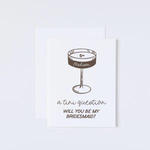 Pode incluir: Um cart&atilde;o branco com uma ilustra&ccedil;&atilde;o marrom de uma ta&ccedil;a de martini com a palavra "Madioon" escrita nela. O texto "a tini question" e "Will you be my bridesmaid?" est&aacute; escrito abaixo da ilustra&ccedil;&atilde;o.