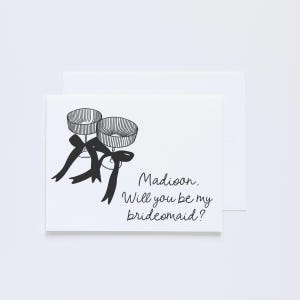 May include: A white card with black ink. The card has a drawing of two champagne flutes tied together with a ribbon. The text on the card reads "Madison. Will you be my bridesmaid?"