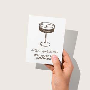 Pode incluir: Um cart&atilde;o branco com um desenho a linha marrom de uma ta&ccedil;a de martini com a palavra "Madison" escrita nela. O texto "a tiny question" e "Will you be my bridesmaid?" est&aacute; escrito abaixo do copo.