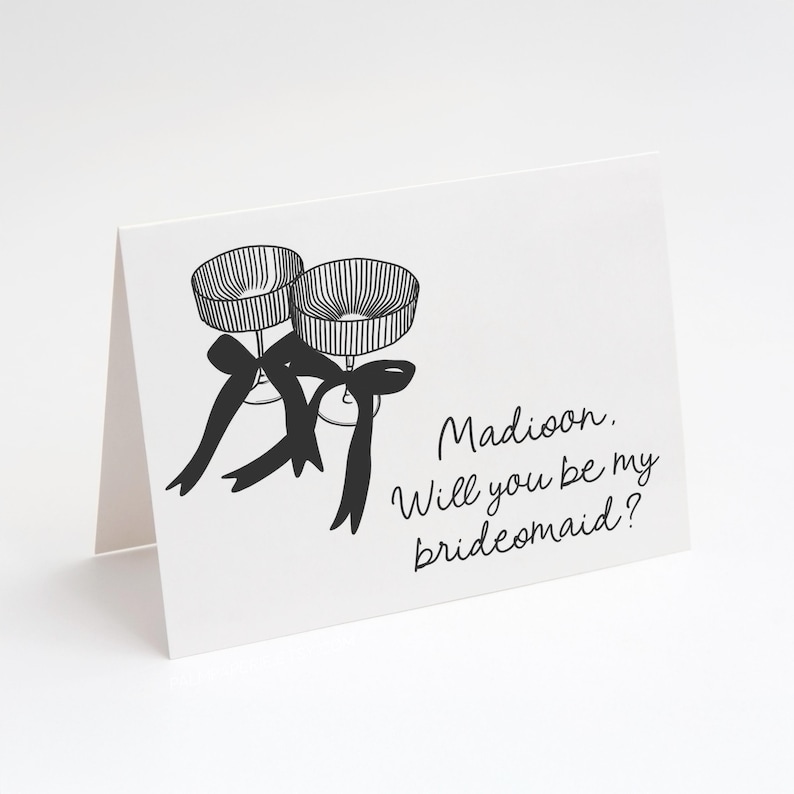 May include: A white card with black ink. Two champagne glasses with black ribbons are drawn on the card. The text "Madison, Will you be my bridesmaid?" is written on the card.