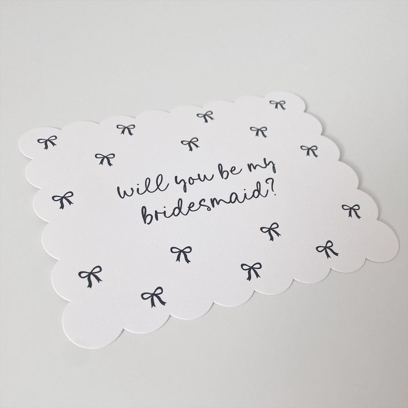 K&ouml;nnte beinhalten: Eine wei&szlig;e Karte mit einem gewellten Rand und dem Text "Will you be my bridesmaid?" in schwarzer Kursivschrift. Die Karte ist mit kleinen schwarzen Schleifen verziert.