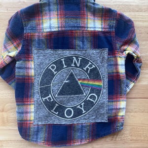 May include: A long-sleeve button-down shirt featuring a blue, red, and yellow plaid pattern. A large, square gray patch is sewn onto the back, displaying the circular Pink Floyd logo with a prism and rainbow spectrum graphic in white ink.