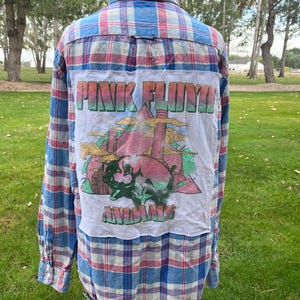 May include: A long-sleeve button-down shirt featuring a blue and red plaid pattern. A white rectangular patch is sewn onto the back, displaying a graphic of a pig with the text PINK FLOYD and ANIMALS in a stylized, multi-colored font.