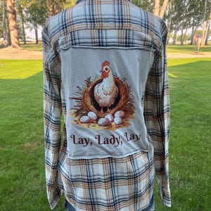 May include: A long-sleeve button-down shirt featuring a tan, white, and navy blue plaid pattern. A rectangular patch is sewn onto the back, displaying a graphic of a white chicken sitting in a nest with eggs and the text Lay, Lady, Lay.