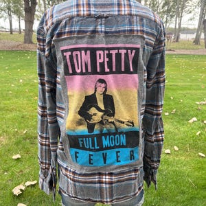 May include: A long-sleeve button-down flannel shirt in a grey, blue, and white plaid pattern. The back features a large rectangular patch with a graphic of Tom Petty holding a guitar, with the text TOM PETTY and FULL MOON FEVER printed.