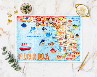 May include: A colorful illustrated jigsaw puzzle of the state of Florida. The puzzle features landmarks, animals, and food items. The puzzle is on a white marble surface with a small potted plant, a gold dish, and a pencil.