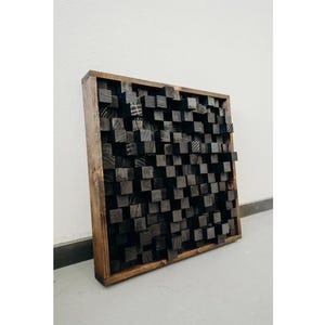 May include: A square wooden wall panel with a dark brown frame and a textured surface made of black wood blocks.