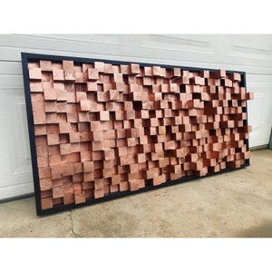 May include: A large wall art piece made of wood blocks in a copper finish. The blocks are arranged in a random pattern and are framed in black.