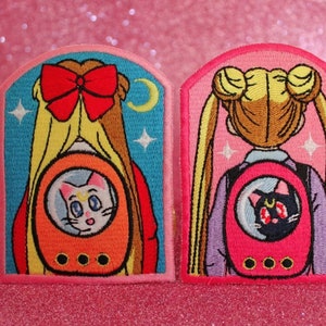 May include: Two embroidered iron-on patches featuring Sailor Moon and her cat Luna. Each patch depicts a character with a backpack and a cat inside a bubble. The patches are pink and blue with white stars.