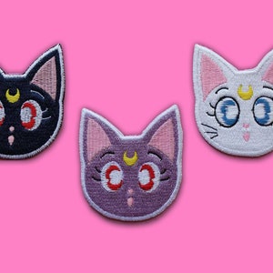Puede incluir: Tres parches bordados para planchar que representan un rostro de gato con una luna creciente y una estrella en la frente. Los parches son negros, blancos y morados.