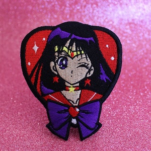 Puede incluir: Parche bordado con un personaje de estilo anime guiñando un ojo, con cabello negro y un atuendo rojo y morado. El parche en forma de corazón tiene un fondo rojo y detalles de estrellas. Una cinta morada en la parte inferior.