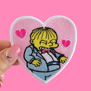 May include: A pink heart-shaped patch with a cartoon character wearing a blue suit and a yellow bow tie. The character has a grumpy expression and there are three pink broken hearts on the patch.