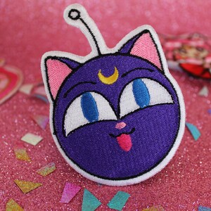 K&ouml;nnte beinhalten: Lila und wei&szlig; gestickter Patch von Luna, der schwarzen Katze aus Sailor Moon, mit einem Halbmond auf der Stirn und herausgestreckter Zunge.