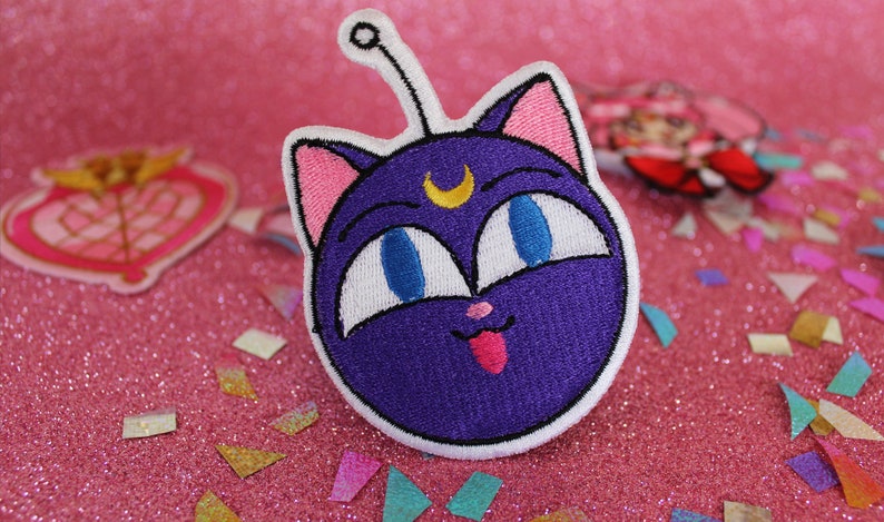 K&ouml;nnte beinhalten: Lila und wei&szlig; gestickter Patch von Luna, der schwarzen Katze aus Sailor Moon, mit einem Halbmond auf der Stirn und herausgestreckter Zunge.