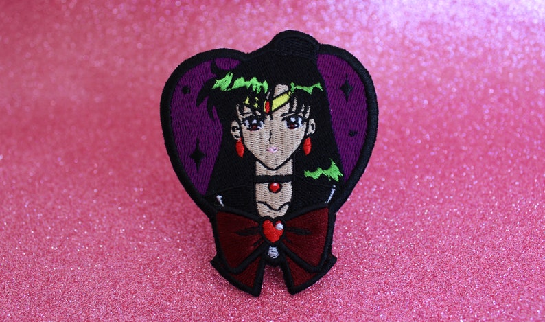 K&ouml;nnte beinhalten: Ein gestickter B&uuml;gel-Patch mit einem violetten Herz, auf dem sich ein Anime-Charakter mit schwarzen Haaren, gr&uuml;nen Augen und einer roten Schleife befindet. Der Charakter tr&auml;gt eine schwarze Halskette mit einem roten Juwel.