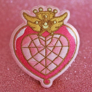 K&ouml;nnte beinhalten: Ein gestickter Patch in Form eines rosa Herzens mit einer goldenen Krone und Fl&uuml;geln. Das Herz hat ein diamantf&ouml;rmiges Design in der Mitte.