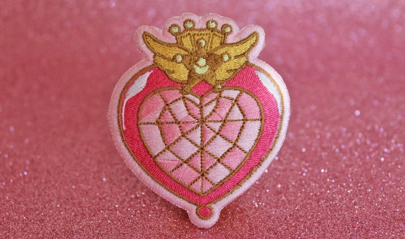 K&ouml;nnte beinhalten: Ein gestickter Patch in Form eines rosa Herzens mit einer goldenen Krone und Fl&uuml;geln. Das Herz hat ein diamantf&ouml;rmiges Design in der Mitte.