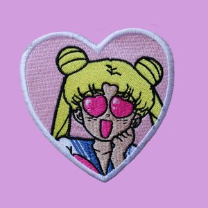 May include: A pink heart-shaped embroidered patch featuring a cartoon character with yellow hair, pink heart-shaped sunglasses, and a surprised expression.