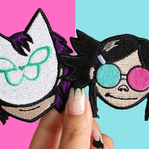 Puede incluir: Dos parches bordados con personajes de dibujos animados. El primer parche es una máscara de gato blanca con alas de mariposa verdes y pelo morado. El segundo parche es un personaje de pelo negro con gafas de sol turquesa y rosas.