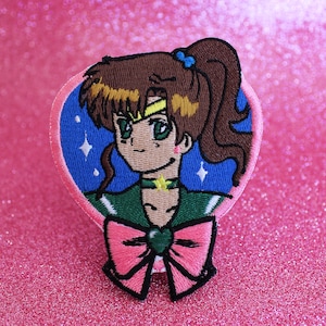 Puede incluir: Un parche bordado para planchar que presenta un personaje de anime de dibujos animados con cabello castaño, ojos verdes y un atuendo verde y azul. El personaje lleva un lazo rosa con una gema verde en el centro. El parche está sobre un fondo de purpurina rosa.