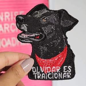 Puede incluir: Parche bordado negro de un perro con un pañuelo rojo con el texto "Olvidar es traicionar" en blanco.